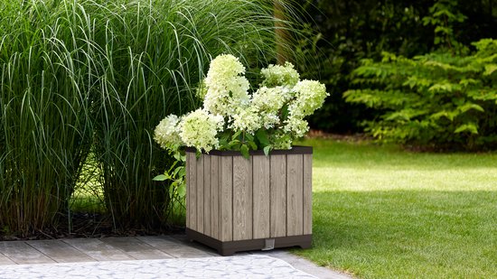 Vierkante grijze plantenbak met houtlook, gevuld met witte hortensia's, in een tuin.