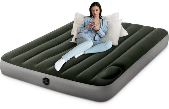 Vrouw zittend op een donkergroen Intex luchtbed met witte kussens, kijkend op haar telefoon.