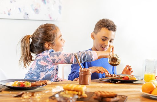Een meisje met een paardenstaart en een jongen met krullend haar delen wafels met stroop en aardbeien aan tafel.