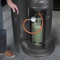 Een persoon opent de klep van een terrasverwarmer om een groene gasfles te tonen, die met een oranje slang is verbonden.