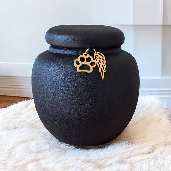 Zwarte mini-urn met gouden pootafdruk en engelenvleugel-bedeltjes. De urn staat op een witte, zachte ondergrond.