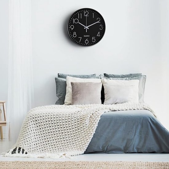 Een zwarte analoge klok hangt aan een witte muur boven een bed met blauw beddengoed en een gebreide deken.