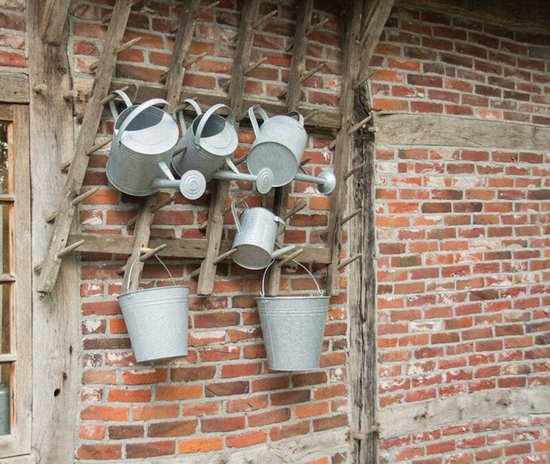 Zinken gieters en emmers hangend aan een houten rek voor een bakstenen muur.