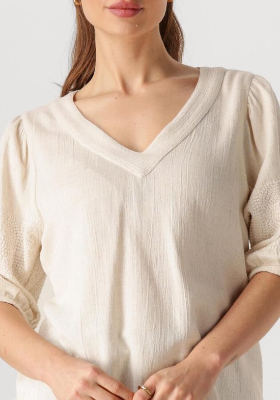 Close-up van een ecru blouse met V-hals en geborduurde details op de kapmouwtjes. Het model raakt de halslijn aan.