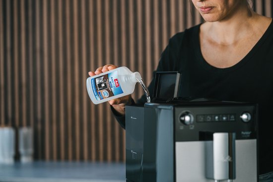 Een vrouw giet Melitta Anti Calc ontkalker uit een fles in een zwarte espressomachine.