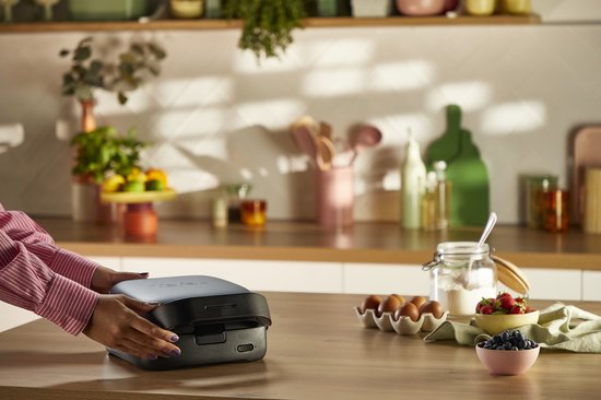 Een vrouw plaatst een gesloten, donkergrijs Tefal wafelijzer op een houten aanrecht.