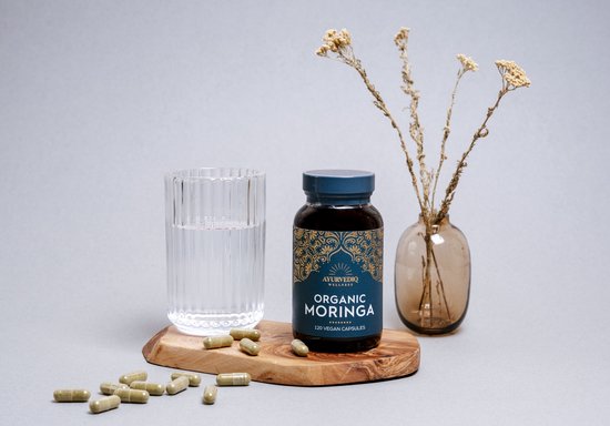 Een bruine fles Ayurvediq biologische moringa capsules naast een glas water en gedroogde takjes op een stuk hout.