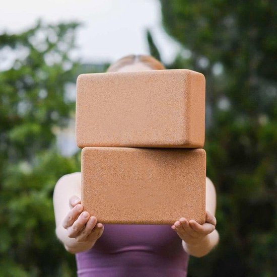 Deux blocs de yoga en liège marron empilés, tenus par une femme portant un haut violet.