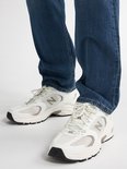 Voeten van een man in een blauwe jeans en witte sneakers. De sneakers hebben het N-logo.