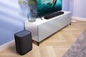 Zwarte Harman Kardon Enchant 900 soundbar op een witte tv-kast, naast een tv en een zwarte subwoofer.