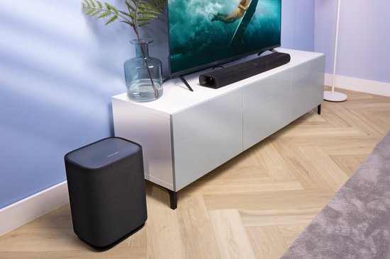 Zwarte Harman Kardon Enchant 900 soundbar op een witte tv-kast, naast een tv en een zwarte subwoofer.