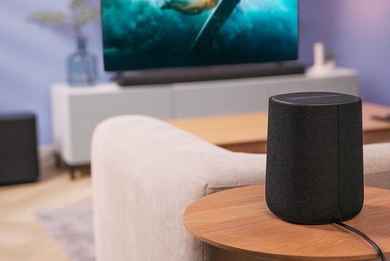 Zwarte Harman Kardon speaker op een houten bijzettafel, met een tv op de achtergrond.
