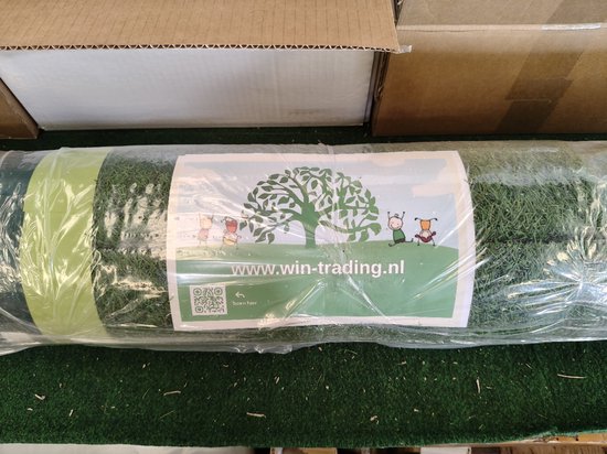 Une pelouse artificielle roulée emballée dans du plastique, avec une étiquette affichant www.win-trading.nl et un arbre avec des personnages.