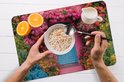 Set de table avec un imprimé d'une maison rose avec une porte bleue, entourée de fleurs, avec des céréales, une orange et du lait.