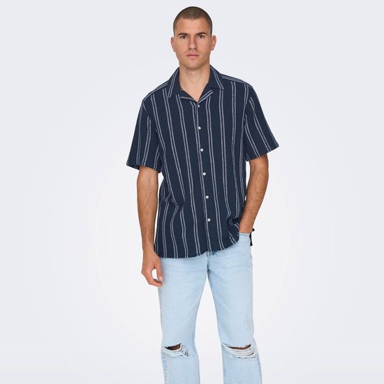 Man met de voorkant naar de camera, draagt een donkerblauw krijtstreeppenshirt en een scheurspijkerbroek.