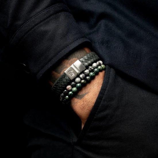 Close-up van een mannelijke arm met drie armbanden: één zwarte gevlochten leren band, één armband met grijze en zwarte kralen, en één met groene kralen.