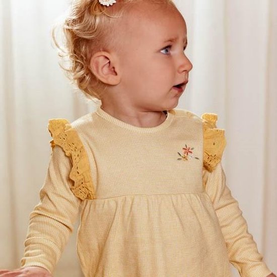 Baby met blonde krulletjes en blauwe ogen draagt een geel geruit jurkje met kanten schouderrandjes.