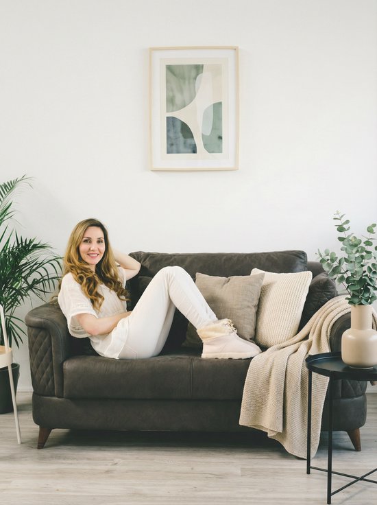 Een vrouw in een witte top en crème broek, zittend op een bank, draagt beige pantoffels.
