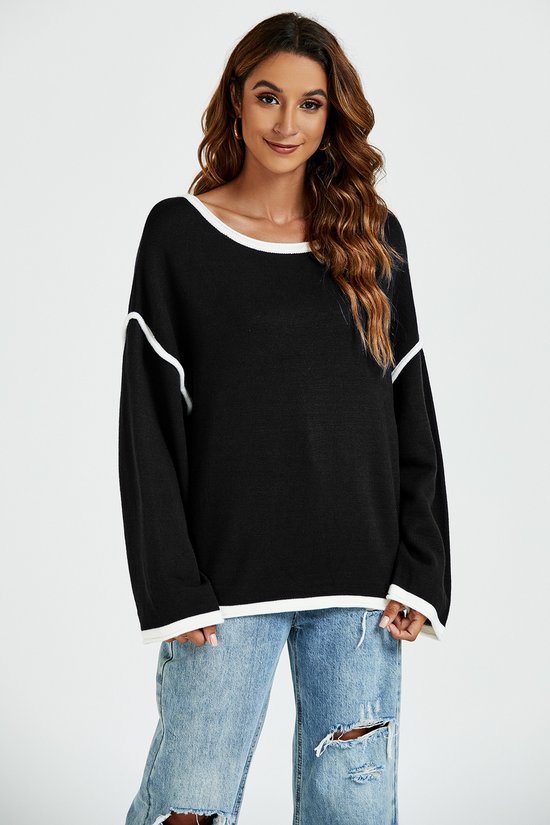 Zwarte trui met witte biezen op de hals, mouwen en zoom. Model staat met een lichte glimlach.