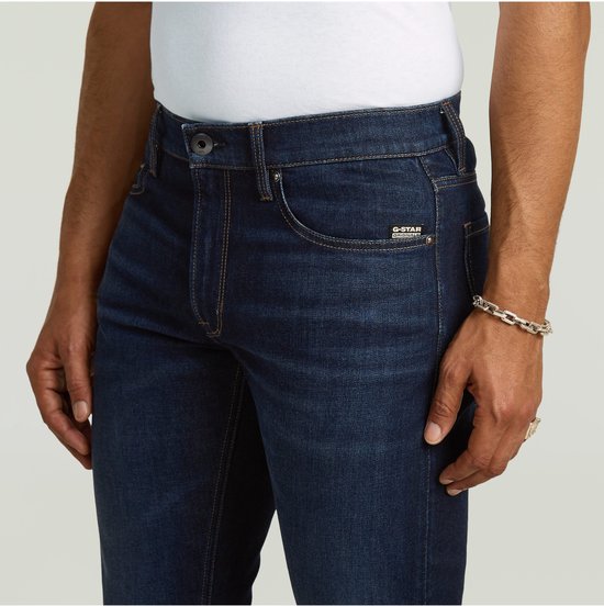 Detail van een donkerblauwe G-Star jeans met G-Star Originals label. Gedragen door een man met een zilveren schakelarmband.