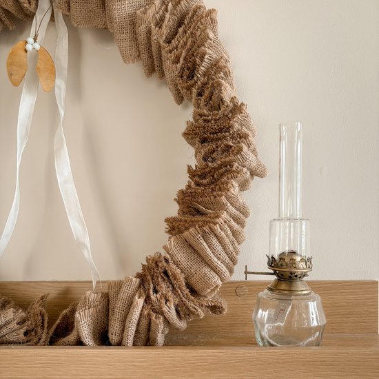 Glazen olielamp met gouden brander, naast een jute krans.