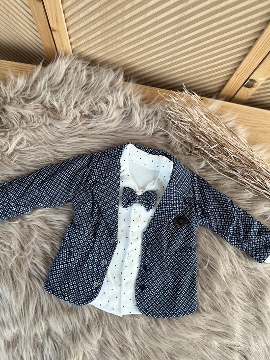 Donkerblauwe blazer met witte print over een wit poloshirt met stippen en strikje.