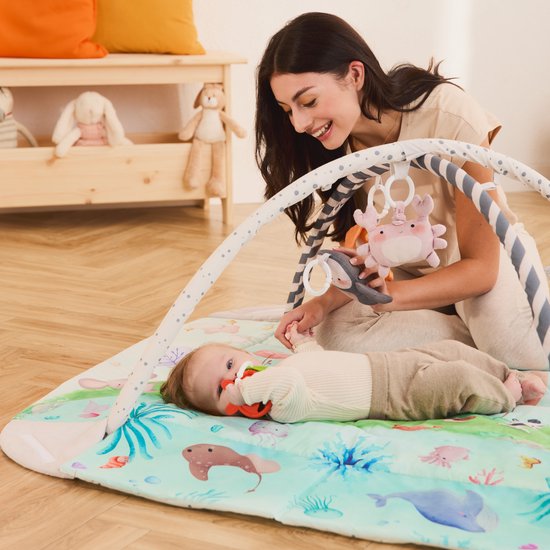 Moeder met baby op een speelkleed met aquarel oceaanmotief, roze krab-speeltje hangt boven.