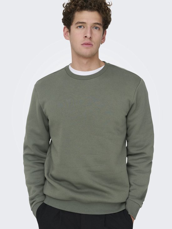Man met handen in de zakken, kijkt naar de camera. Draagt een effen groene sweater met een wit t-shirt eronder.