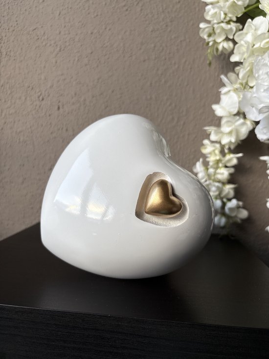 Wit hartvormige urn met een gouden hartje, naast witte bloemen.