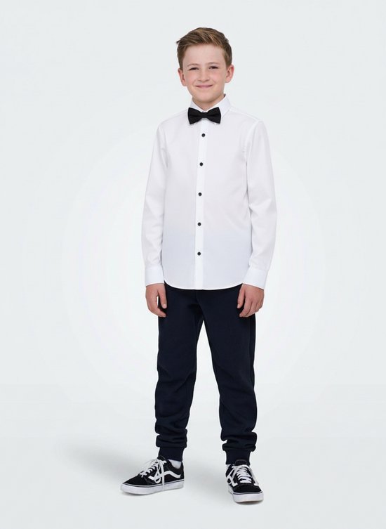 Jongen in wit overhemd met zwarte vlinderdas en zwarte knopen, staand met zwarte broek en sneakers.