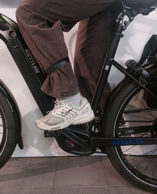 Een persoon op een fiets, met bruine broek en witte schoenen. Een zwarte broekklem met 'RIEBA'-logo is om de broekspijp bevestigd.