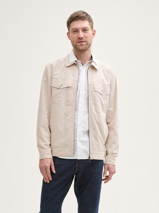 Man met beige linnen jack met ritssluiting, twee borstzakjes met knoop, gedragen met een wit overhemd en spijkerbroek.