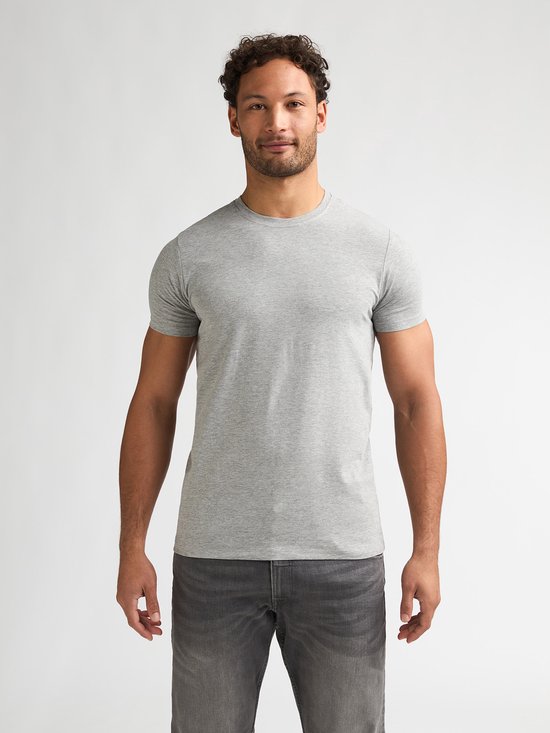 Man met krullend haar draagt een effen lichtgrijs T-shirt met ronde hals en grijze jeans.