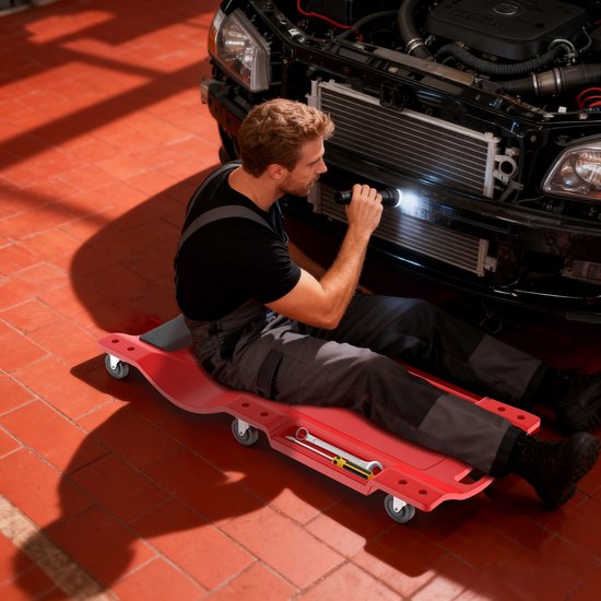 Man in overall op rode rolplank, gebruikt zaklamp om aan de motor van een auto te werken.