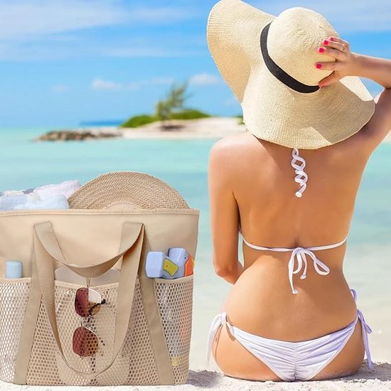 Un sac de plage beige avec poches en filet, rempli d'articles de plage, est posé sur le sable à côté d'une femme en bikini regardant l'océan.