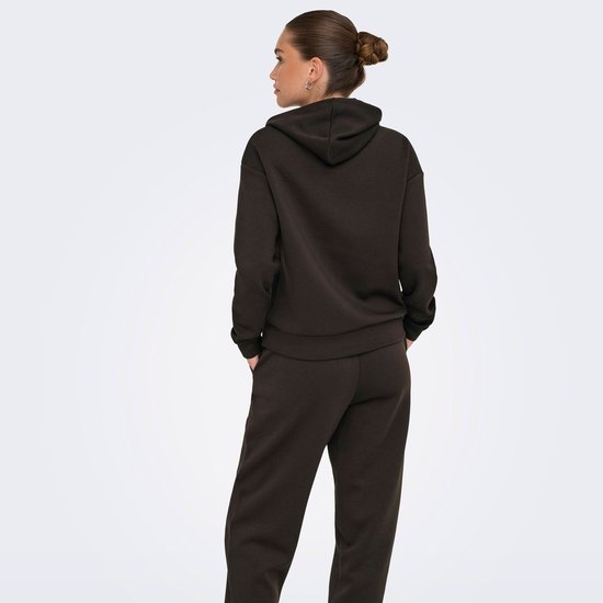Model staat met de rug naar de camera, handen in de zakken, in een donkerbruine hoodie.