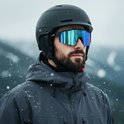 Man met baard, draagt een zwarte skihelm en een KAPVOE sportbril met spiegelende blauw-groene lenzen tijdens sneeuwval.