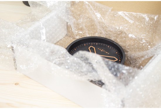 Een zwart, rond horloge met koperkleurige wijzers, verpakt in noppenfolie in een kartonnen doos.
