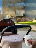 Zwarte ZAZU Baby Rocker aan een duwstang van een beige kinderwagen, met een hand die een koffiebeker vasthoudt.