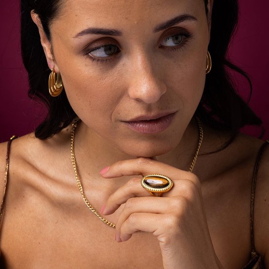 Close-up van een model die een gouden ring met een ovaal tijgeroog en gouden oorringen draagt. De ring is een statement.