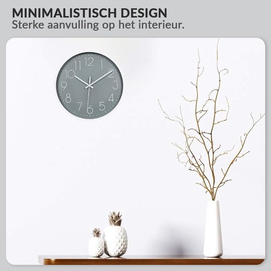 Minimalistische grijze wandklok met witte wijzers en cijfers, hangend aan een muur boven een plank met decoratie.