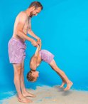 Man houdt jongen bij de enkels, die ondersteboven hangt in paarse zwembroeken met cirkelpatroon.