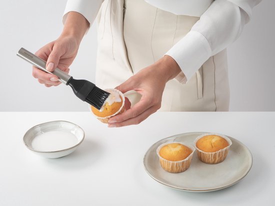 Mains glaçant un cupcake avec un petit pinceau de pâtisserie argenté à poils noirs.
