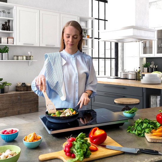 Vrouw kookt groenten in een pan op een zwarte inductie kookplaat in een moderne keuken.