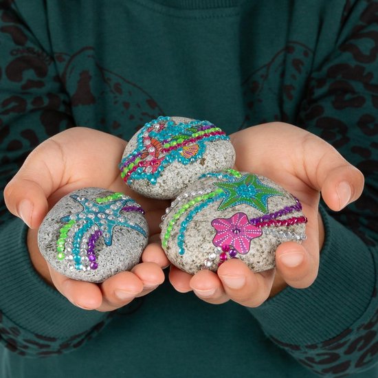 Kinderhanden houden drie grijze stenen vast, versierd met strass steentjes, zeesterren en bloemen.