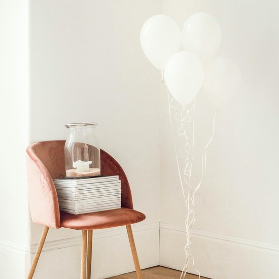 Roze fluwelen stoel met houten poten, met een stapel tijdschriften en een glazen vaas erop. Drie witte ballonnen gebundeld ernaast.