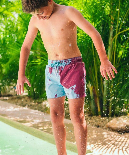 Jongetje in lichtblauwe zwembroek met paarse verfvlekken, spattend bij een zwembad, met groene planten op achtergrond.