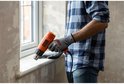 Man met blauw geruit shirt en grijze handschoenen gebruikt een oranje Black+Decker verfbrander bij een raamkozijn.