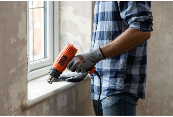 Man met blauw geruit shirt en grijze handschoenen gebruikt een oranje Black+Decker verfbrander bij een raamkozijn.