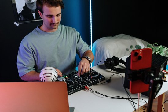 Man met snor en blauw T-shirt bedient een zwarte DJ-controller met knoppen en draaitafels, met een witte microfoon en koptelefoon ernaast.